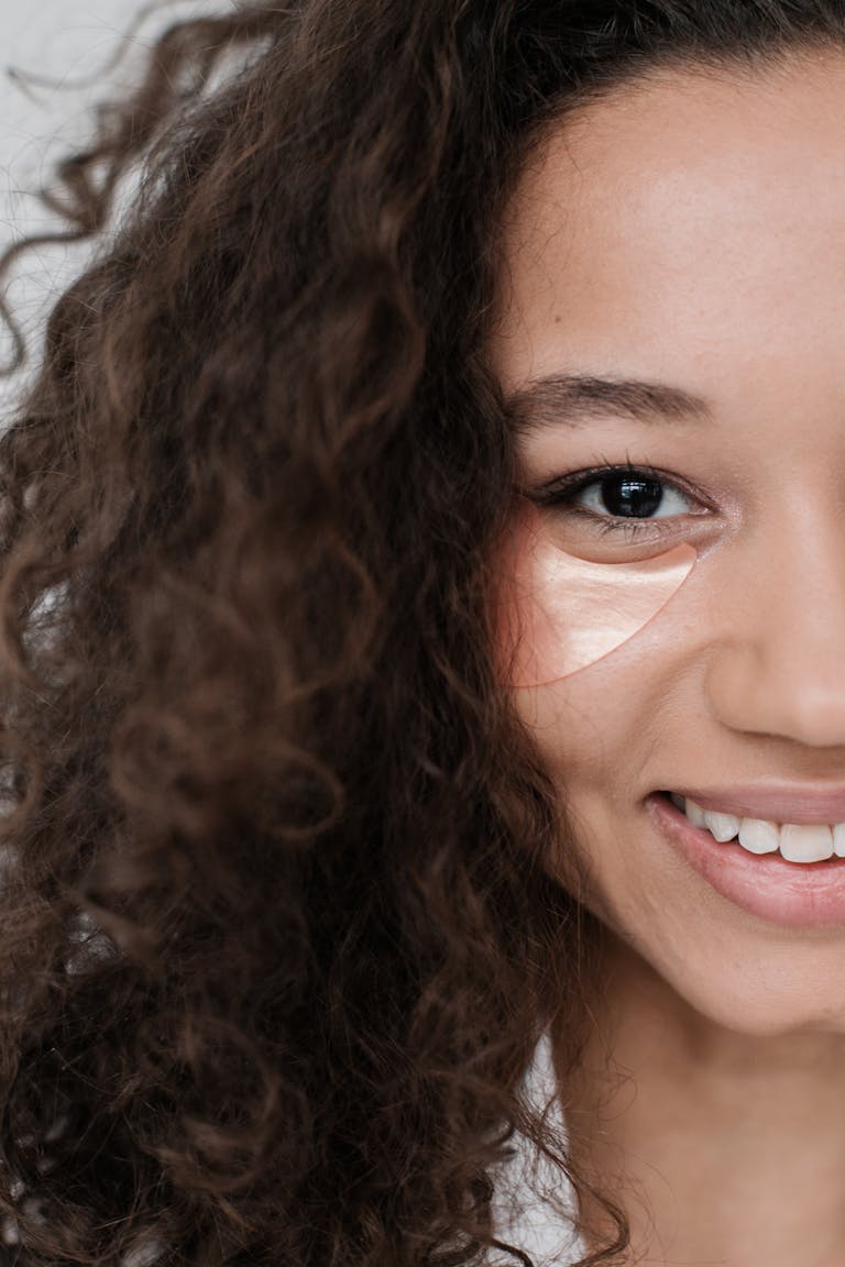 Close-up of a smiling woman with curly hair using an eye patch for skincare.